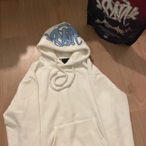 Vit hoodie från Stay Sane med blått tryck - Säljer en vit hoodie från Stay Sane i storlek S. Tröjan har en stor huva med blått graffiti-tryck och grova snören. Klassisk känguruficka framtill och mjukt material som känns skönt mot huden. Perfekt för dig som gillar streetwear och vill sticka ut.