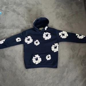 Snygg mörktblå hoodie  - Säljer en marinblå hoodie från Denim Tears med unika vita blommönster över hela tröjan. Tröjan har huva, magficka och är tillverkad i ett mjukt material som känns skönt mot huden. Perfekt för dig som vill sticka ut med en cool och kreativ stil.