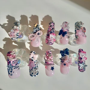  rosa och svart press-on naglar - Set med 10 press-on naglar i rosa och svart med zebramönster, pärlor, stjärnor, rosetter och Hello Kitty-dekorationer. Naglarna har 3D-detaljer i silver, glitter och pärlemor, samt mix av matta och glansiga ytor. Perfekt för dig som älskar maximalistisk och kawaii stil.