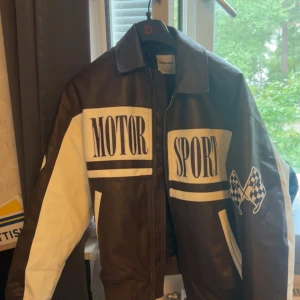 Brun och vit skinnjacka Bershka - Cool brun och vit skinnjacka från Bershka med stora MOTOR SPORT-tryck och rutiga flaggor på framsidan. Jackan har dragkedja, klassisk krage och kontrasterande vita detaljer på ärmar och fickor. Perfekt för dig som gillar motorsport vibes. Oversized