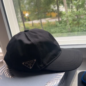Svart keps från Prada - Snygg svart keps från Prada med klassisk böjd skärm och ikonisk metalltriangel med logga på sidan. Kepsen har stilren design och är tillverkad i ett slätt syntetmaterial som ger en exklusiv känsla. Perfekt för dig som gillar designeraccessoarer.