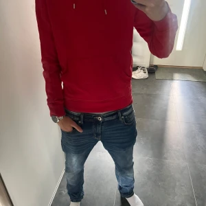 Röd hoodie från J.Lindeberg - Säljer en snygg röd hoodie från J.Lindeberg med huva och snörning. Tröjan har en klassisk känguruficka framtill och långa ärmar. Perfekt för dig som gillar stilrena och bekväma plagg. Passar dig som vill ha en enkel men cool look.