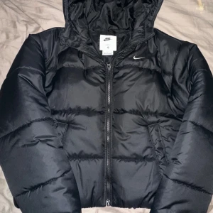 Svart pufferjacka från Nike Therma-Fit - Svart pufferjacka från Nike med Therma-Fit-teknologi. Jackan har huva, dragkedja framtill och diskret Nike-logga på bröstet. Fylld för extra värme och har en clean, sportig look. Perfekt för kalla dagar när du vill hålla stilen.