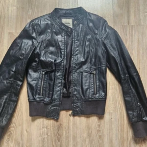 Svart skinnjacka från Bershka  - Snygg svart skinnjacka från Bershka i storlek S. Jackan har dragkedja framtill, två fickor med dragkedjor och ribbade muddar vid ärmslut och midja. Klassisk bomberstil med rund krage och coola sömdetaljer. Perfekt för dig som vill ha en edgy look.