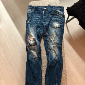 Blå Dsquared2 jeans med slitningar - Säljer ett par blå jeans från Dsquared2 med coola slitningar, patchar och detaljerade sömmar. Jeansen har en klassisk femficksdesign, tydliga slitna partier och en snygg Dsquared2-label bak. Passar dig som gillar street och edgy stil.