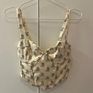 Beige korsett-topp  - Superfin beige korsett-topp med svarta cowboy boots-mönster över hela tyget. Toppen har breda axelband, markerade sömmar och smockad rygg. Använd en gång.