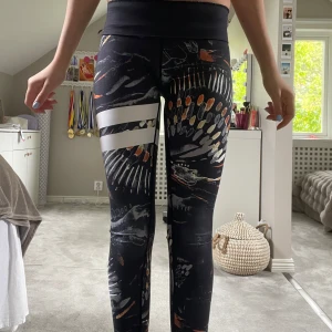 Stronger leggings med mönster XXS - Snygga leggings från Stronger i svart med färgglatt abstrakt mönster och två vita ränder på ena benet. Byxorna är ner vikta på de två första bilderna. Mid waist/ low waist om man viker ner. Materialet är 73% polyester och 27% elastan för maximal stretch och komfort. 