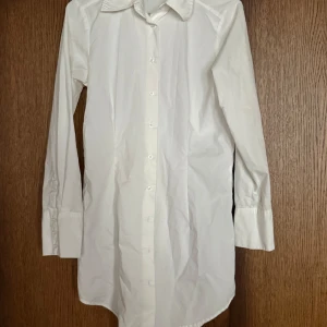 Vit långärmad skjorta med knappar - Klassisk vit skjorta i bomull med lång ärm och knappar hela vägen framtill. Lång modell. Skjortan har bred krage och manschetter med tre knappar. Perfekt att styla med jeans eller kjol för en clean look.