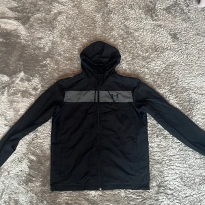 Svart vindjacka från Under Armour - Svart vindjacka från Under Armour med grått band över bröstet och logga. Jackan har huva, hel dragkedja och fickor med dragkedja.