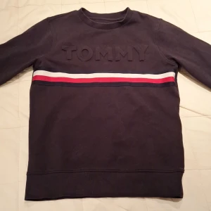 Mörkblå Tommy Hilfiger sweatshirt - Snygg mörkblå sweatshirt från Tommy Hilfiger med upphöjd TOMMY-text på bröstet och klassiska ränder i vitt, rött och blått över bröstet. Långärmad modell med ribbade muddar och liten logga på ärmen. Perfekt för en chill och stilren look.
