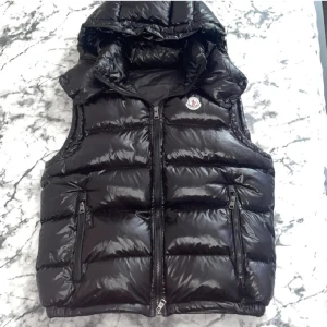 Svart dunväst från Moncler - Efterlängtad tröja från monclear som jag tyvärr behöver sälja nu, använd 1-3 gånger och inga tecken på skador. Snygg svart dunväst från Moncler med glansig finish och huva. Västen har två dragkedjefickor framtill, Moncler-logga på bröstet och quiltad design. Perfekt för lager-på-lager och håller dig varm under kyliga dagar. Perfekt för dig som vill ha lite stil.