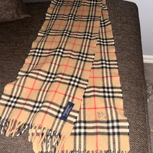 Beige rutig halsduk från Burberry - Snygg beige halsduk från Burberry med klassiskt rutigt mönster i svart, vitt och rött. Halsduken är lång med fransar i ändarna och har en broderad Burberry-logga. Tillverkad i mjuk ull som känns lyxig och varm. Perfekt accessoar för att lyfta din outfit. Köpt i london