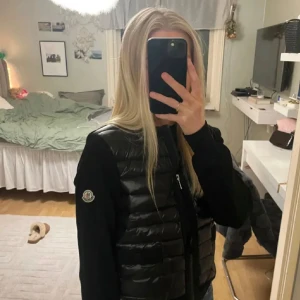 Svart dunväst från Moncler - Snygg svart dunväst från Moncler med quiltad framsida och dragkedja. Västen har rund halsringning, två fickor framtill och Moncler-logga på ärmen. Materialmix med dun och stickade partier ger en cool och sportig look.