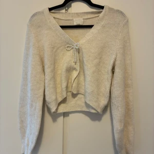 Beige/vit stickad kofta med pärlrosett - Superfin beige/vit stickad kofta med croppad passform och lång ärm. Koftan har en v-ringning och stängs framtill med en söt rosett av pärlor. Perfekt för lager-på-lager och ger en mjuk, trendig look.