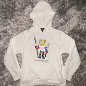 Vit hoodie Polo Bear Ralph Lauren - Vit hoodie från Polo Ralph Lauren med ikoniskt Polo Bear-tryck framtill. Tröjan har huva, långa ärmar och är gjord i mjuk bomull. Perfekt för dig som gillar streetstyle och vill sticka ut med en cool björn på bröstet. Kom privat för mer info och pris!💯