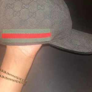 Svart Gucci keps med GG-mönster - Snygg svart keps från Gucci med klassiskt GG-monogram över hela kepsen och ikoniska gröna och röda ränder på sidan. Kepsen är tillverkad i bomull och har en böjd skärm. Perfekt för dig som vill ha en lyxig och stilren accessoar.