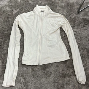 Vit ziptröja med lång ärm - Säljer en vit långärmad ziptröja med hög krage och figurnära passform. Tröjan har dragkedja hela vägen och tumhål vid ärmsluten, vilket ger en sportig och clean look. Perfekt för lager-på-lager eller till träning.