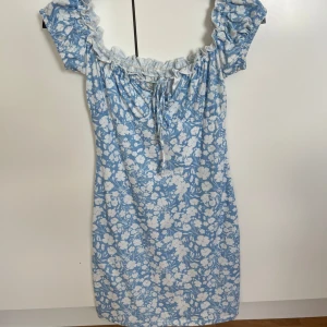 Blommig blå kortklänning - Supersöt blå kortklänning med vitt blommönster och volang vid axlarna. Använd 1 gång. Mellan XS och S i storlek.