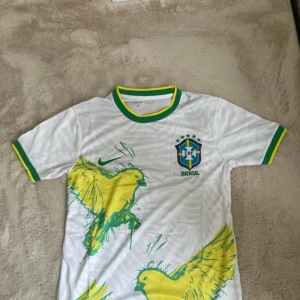 Vit Brasilien fotbollströja Nike - Vit Brasilien fotbollströja från Nike med gröna och gula detaljer på krage och ärmslut. Framsidan har stora gula fåglar och CBF-logga på bröstet. Tillverkad i Dri-FIT-material som andas, perfekt för träning eller match.
