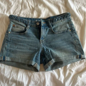 Blå jeansshorts med flätad detalj - Snygga blå jeansshorts från You med flätad detalj i midjan och klassisk femficksdesign. Shortsen har uppvikta benslut och stängs med knapp och dragkedja. Perfekta för varma dagar och ger en avslappnad vibe.