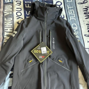 Svart Sail Racing Gore-Tex orca jacket - Grym svart Sail Racing Orca Jacket med Gore-Tex-material och schyssta detaljer. Jackan har hög krage, dragkedja framtill, flera fickor och coola loggor på ärmarna. Perfekt för dig som vill ha en teknisk och stilren vindjacka med sportig känsla. Nypris på denna ligger på 12000kr. endast testad en gång. priset ej hugget i sten vid snabbaffär!