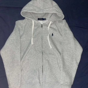 Grå hoodie från Polo Ralph Lauren - Snygg ljusgrå hoodie från Polo Ralph Lauren med dragkedja, vit snörning i huvan och klassisk broderad logga på bröstet. Mjuk bomullsblandning och känguruficka framtill. Perfekt för chill dagar eller när du vill ha en clean look. Tröjan är oanvänd med pris lapp.