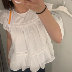 Vit volangblus med spetsdetaljer - Supersöt vit blus med volangärmar och flera lager av tunna volanger. Blusen har fina spetsdetaljer och är tillverkad i ett lätt, luftigt material som känns perfekt för varmare dagar. Den har en lös passform och är ärmlös med romantisk känsla.