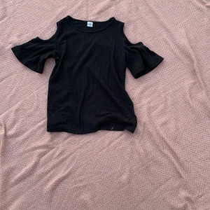 Svart ribbad topp med cut out från Lindex - Svart ribbad topp från Lindex med trendiga cut out-detaljer på axlarna och korta, utsvängda ärmar. Toppen har en enkel rund halsringning och är tillverkad i mjukt bomullsmaterial. Perfekt för dig som gillar en stilren men edgy look.