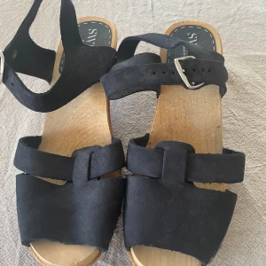 Svarta sandaler från Sweeks i mocka - Svarta träskor från Sweeks med breda remmar i mocka och justerbart spänne runt ankeln. Sulan är i trä med räfflad gummiyta undertill för extra grepp. Perfekta för en chill sommarlook.