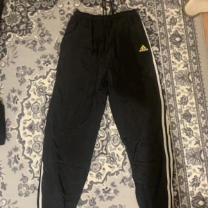 Svarta Adidas träningsbyxor med ränder - Svarta träningsbyxor från Adidas med klassiska vita ränder längs sidorna och gul logga på låret. Byxorna har resår och snörning i midjan, dragkedja vid benslut och är fodrade med vitt tyg. Perfekta för sport eller chill.