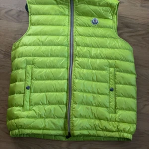 Neongul dunväst från Moncler - Säljer en riktigt snygg neongul dunväst från Moncler. Västen är quiltad med horisontella sömmar, har dragkedja framtill och två fickor med tryckknappar. Perfekt för lager-på-lager och sticker verkligen ut i mängden.