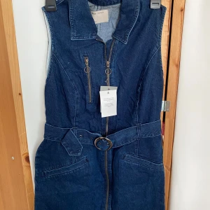 Ärmlös jeansklänning från ASOS - Mörkblå ärmlös jeansklänning från ASOS med markerad krage, dragkedjor framtill och bälte i midjan. Klänningen har fickor och coola metalldetaljer som ger en edgy vibe. Perfekt för dig som gillar denim och vill sticka ut. Strl 40/42 lappen finns kvar o den är inte använd.