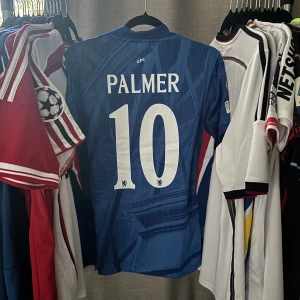 Chelsea Palmer 10 Champions League fotbollströja - Chelsea FC matchtröja med Palmer 10-tryck, Champions League-märke på ärmen och klubbmärke på bröstet. Snyggt geometriskt mönster i olika blå nyanser, klassisk krage och korta ärmar. Perfekt för dig som vill sticka ut på läktaren eller i stan.