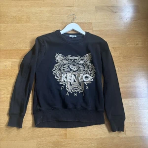 Svart Kenzo tröja med tigerbrodyr - Svart tröja från Kenzo med ikonisk tigerbrodyr och texten 'KENZO PARIS' framtill. Klassisk rund halsringning och långa ärmar. Tillverkad i mjuk bomull, perfekt för en cool och avslappnad stil.