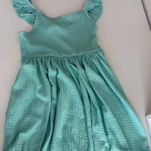 Turkos kortklänning med volangärmar - Supersöt turkos barn kortklänning med smockad struktur och volangdetaljer på axelbanden. Klänningen har en luftig, rynkad kjol och är ärmlös med breda axelband. Perfekt för varma dagar och ger en fräsch sommarkänsla. Ord pris 229kr. Pris kan diskuteras.