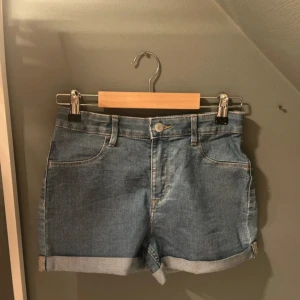 Blå jeansshorts med upprullad kant - Klassiska blå jeansshorts med upprullad kant och femficksmodell. De har bälteshällor, knapp och dragkedja framtill samt bakfickor. Perfekta för varma dagar och enkel att matcha med allt.