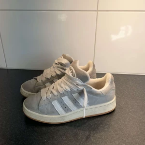Adidas Campus grå sneakers mocka - Snygga Adidas Campus sneakers i ljusgrå mocka med klassiska vita ränder på sidorna och vit text. Skorna har vit sula med gummidetalj och breda vita skosnören. Perfekta för dig som gillar en clean och stilren look. Har inte försökt rengöra dom men dom kommer inte till användning längre. 