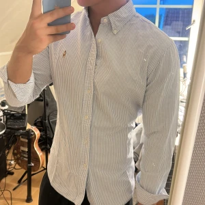 Randig skjorta från Polo Ralph Lauren - Säljer en klassisk skjorta från Polo Ralph Lauren i vitt och blått med tunna vertikala ränder. Skjortan har button-down krage, broderad logga på bröstet och långa ärmar. Tillverkad i mjuk bomull och har en normal passform. Perfekt för en clean och stilren look. En liten defekt vid kragen inget som syns när den e på!