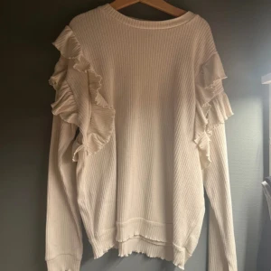 Vit ribbad tröja med volang från Kiabi - Supersöt vit ribbad tröja från Kiabi med långa ärmar och volangdetaljer längs axlarna. Tröjan har en mjuk känsla och är perfekt för dig som gillar en stilren men ändå lekfull look. Passar till både jeans och kjol.