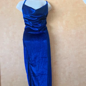 Blå sammetsklänning med slits - Snygg långklänning i djupblå sammet med smala axelband och v-ringad urringning. Klänningen har en elegant slits framtill som ger extra edge. Perfekt för dig som vill sticka ut med en lyxig och trendig look.