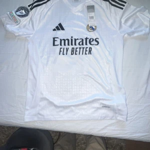 Real Madrid vit matchtröja Adidas - Snygg vit Real Madrid fotbollströja från Adidas med svarta detaljer och klubbmärke på bröstet. Tröjan har tryck med 'Emirates Fly Better' samt nummer 12 och Trent på ryggen. Helt ny inte ens använd en gång. Billigt. Storleken är M men den passar perfekt för personer med S storlek 