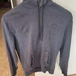 Hugo boss grå tröja - Snygg grå hoodie från Hugo BOSS med broderad BOSS-logga på bröstet. Tröjan har huva med snörning och en stor magficka framtill. Perfekt för en chill och stilren look. Materialet känns mjukt och skönt mot huden. Nypris 1000kr