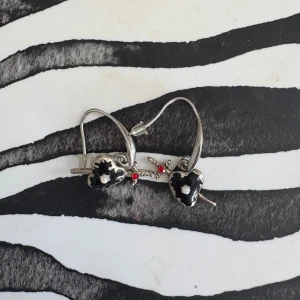 Silvriga örhängen med svarta hjärtan - Unika örhängen i silverfärg med svarta hjärtan och små vita detaljer. Varje hjärta är genomborrat av ett svärd och dekorerat med en röd sten. Perfekt accessoar för dig som gillar edgy och cool stil.