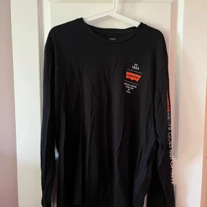 Svart långärmad Levis tröja - Svart långärmad tröja från Levis med tryckt logga och text på bröstet samt tryck längs ena ärmen. Tröjan har en klassisk rund halsringning och är gjord i mjuk bomull. Perfekt för en avslappnad och stilren look.