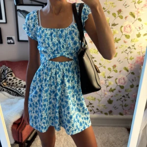 Blå blommig klänning  - Supersöt kortklänning med blått blommönster. Klänningen har volangärmar, fyrkantig ringning och en snygg cut-out detalj i midjan. Det ser ut som en klänning men igentligen är det shorts som är perfekt för blåsigt väder