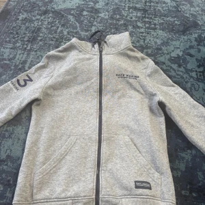 Grå hoodie från Race Marine XS - Säljer en grå hoodie från Race Marine i storlek XS. Tröjan har dragkedja framtill, huva med snörning och två fickor. Det finns tryck på ena ärmen och bröstet. Perfekt för dig som gillar sportig och avslappnad stil.