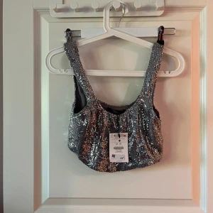 Glittrig silver topp med paljetter - Säljer en glittrig silver topp från Cider, täckt med paljetter som verkligen fångar ljuset. Modellen är croppad med breda axelband och en rundad halsringning. Perfekt för dig som vill sticka ut och glänsa på nästa fest!