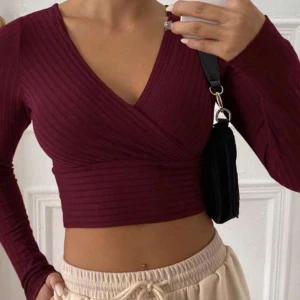 Ribbad omlottopp i vinrött och svart - Säljer två croppade omlottoppar med ribbad struktur, en i vinrött och en i svart. Båda har lång ärm och djup v-ringning, perfekta att styla med höga byxor eller kjol. Mjukt och stretchigt material som sitter snyggt på kroppen.