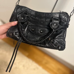 Svart axelväska i skinnstil - Cool svart axelväska med flätat handtag, dragkedja och snygga metalldetaljer. Väskan har både axelrem och handtag, samt en liten ficka framtill med dragkedja. Perfekt storlek för det viktigaste och ger en edgy vibe till din outfit.