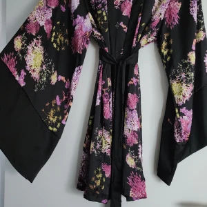 Svart blommig kimono-klänning M/L - Svart kimono-inspirerad klänning med stora vida ärmar och knytband i midjan. Kimonon har ett färgstarkt blommönster i rosa, lila och gult. Perfekt för dig som vill sticka ut med en unik och flowy look. Endast testad
